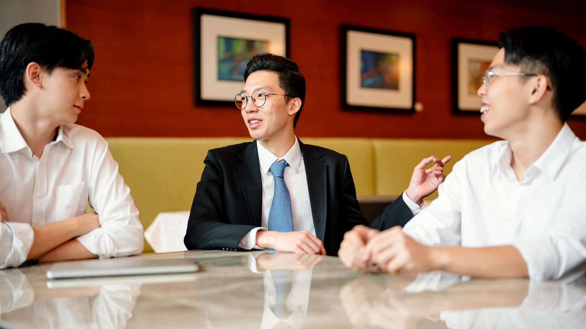 Taylor's students sit around a table in a restaurant or hospitality setting, engaged in discussion, with one student in formal business attire speaking while the others listen and smile.