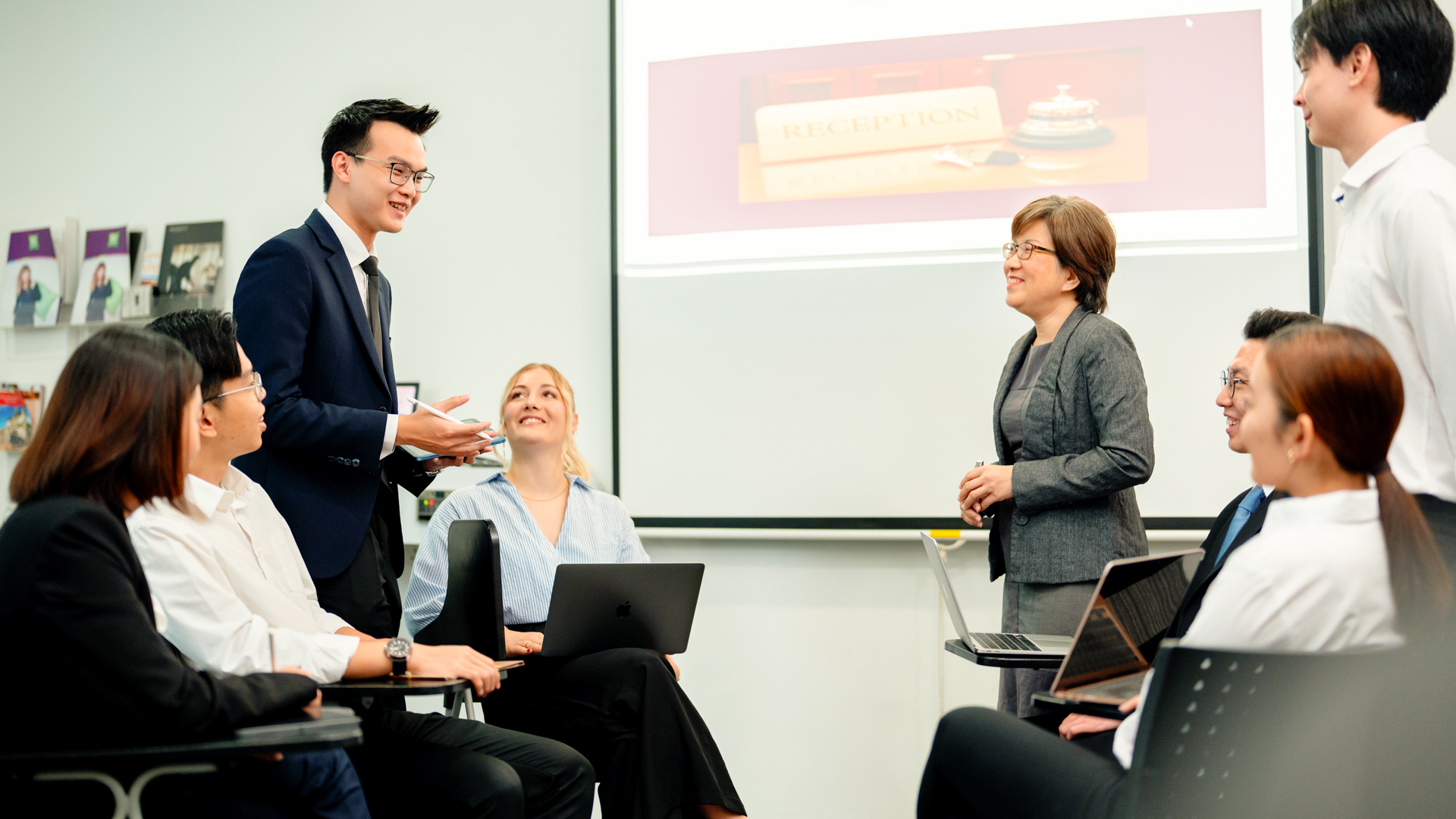 A group of Taylor's students and a lecturer participate in a classroom discussion, with several people using laptops while a presentation about hospitality or tourism is displayed on a screen in the background.