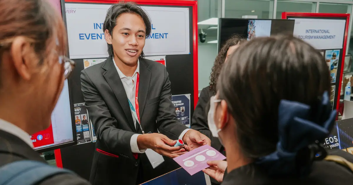 A Taylor's student stands at an exhibition booth, handing a brochure or information card to visitors while discussing a project, with presentation displays visible in the background.