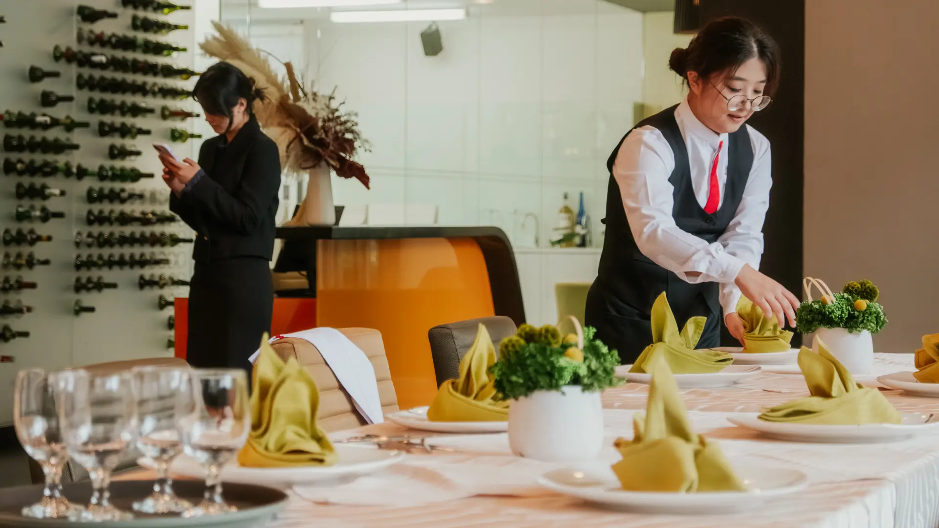 A Taylor's student arranges table settings at a formally prepared dining table, while another student stands nearby using a phone, in a restaurant training or event management environment.