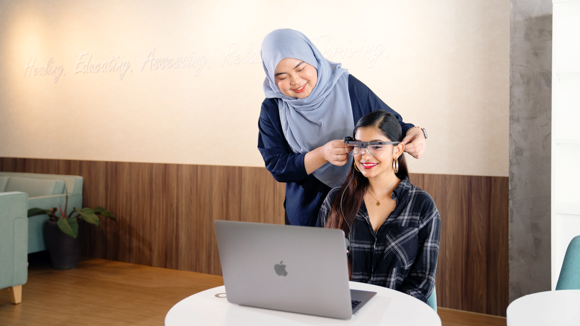 A lecturer fits specialized glasses on another seated at a laptop in a clinical or research setting.