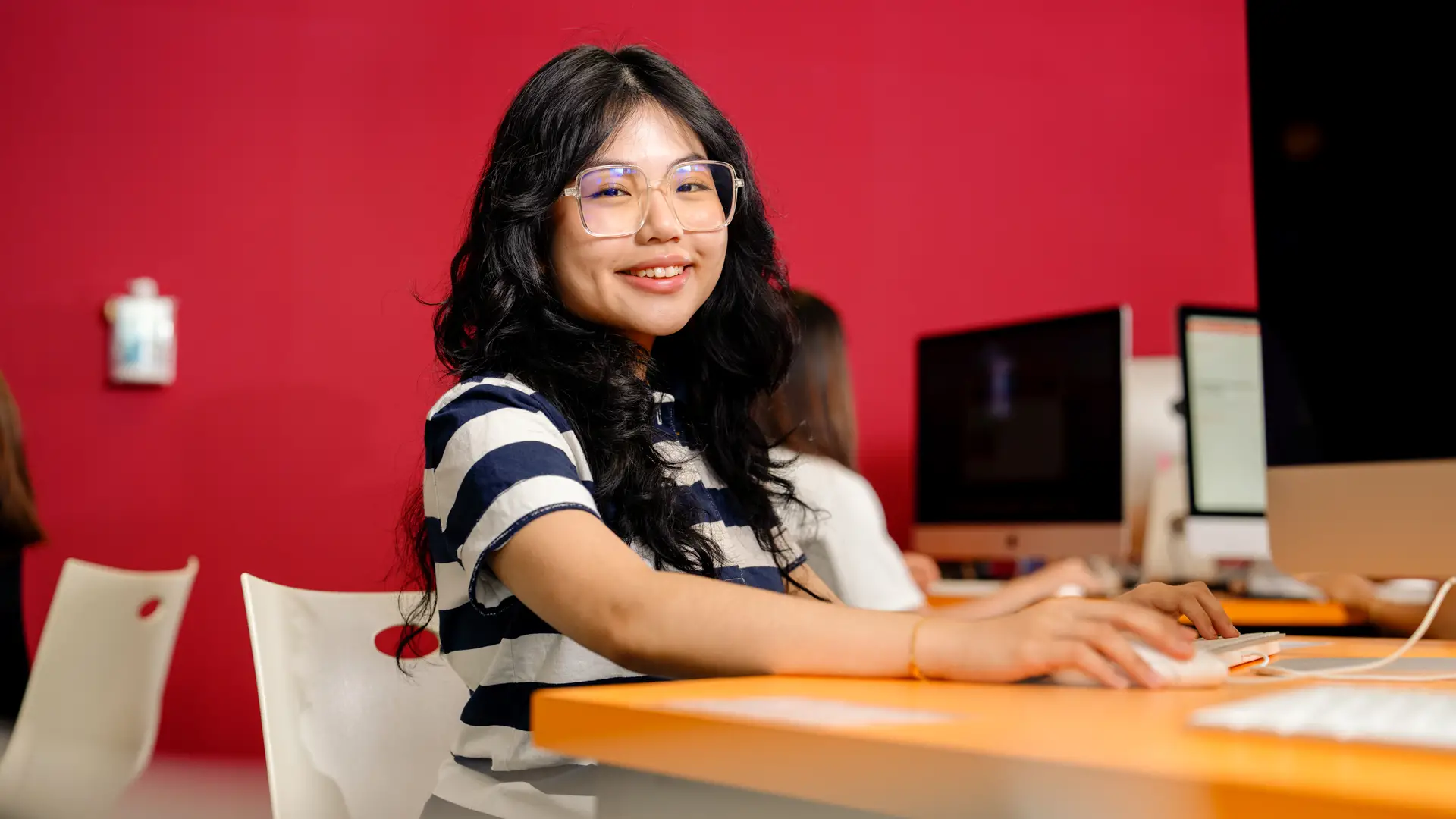 A Taylor's student wearing glasses smiles while using a computer in a classroom.