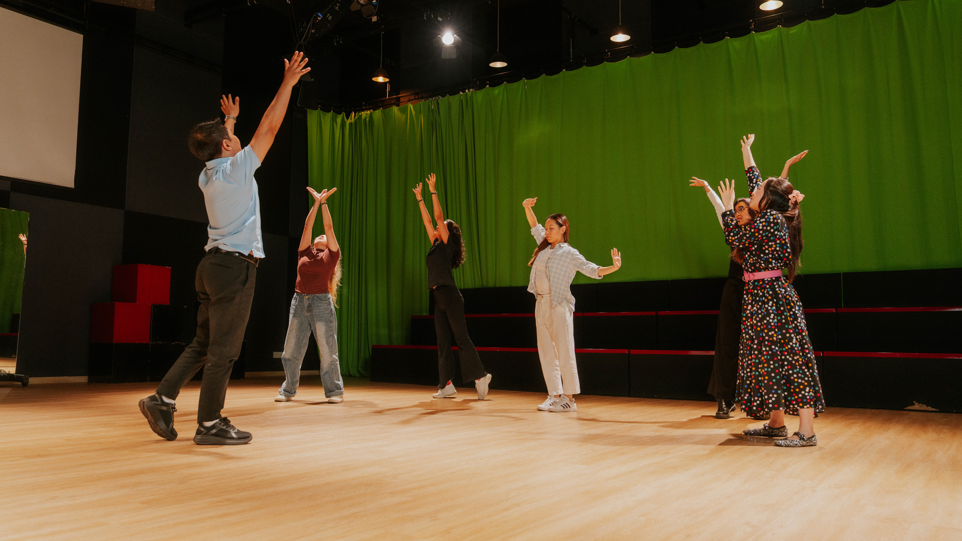 A group of Taylor's students practice expressive movements on a stage with a green curtain backdrop.