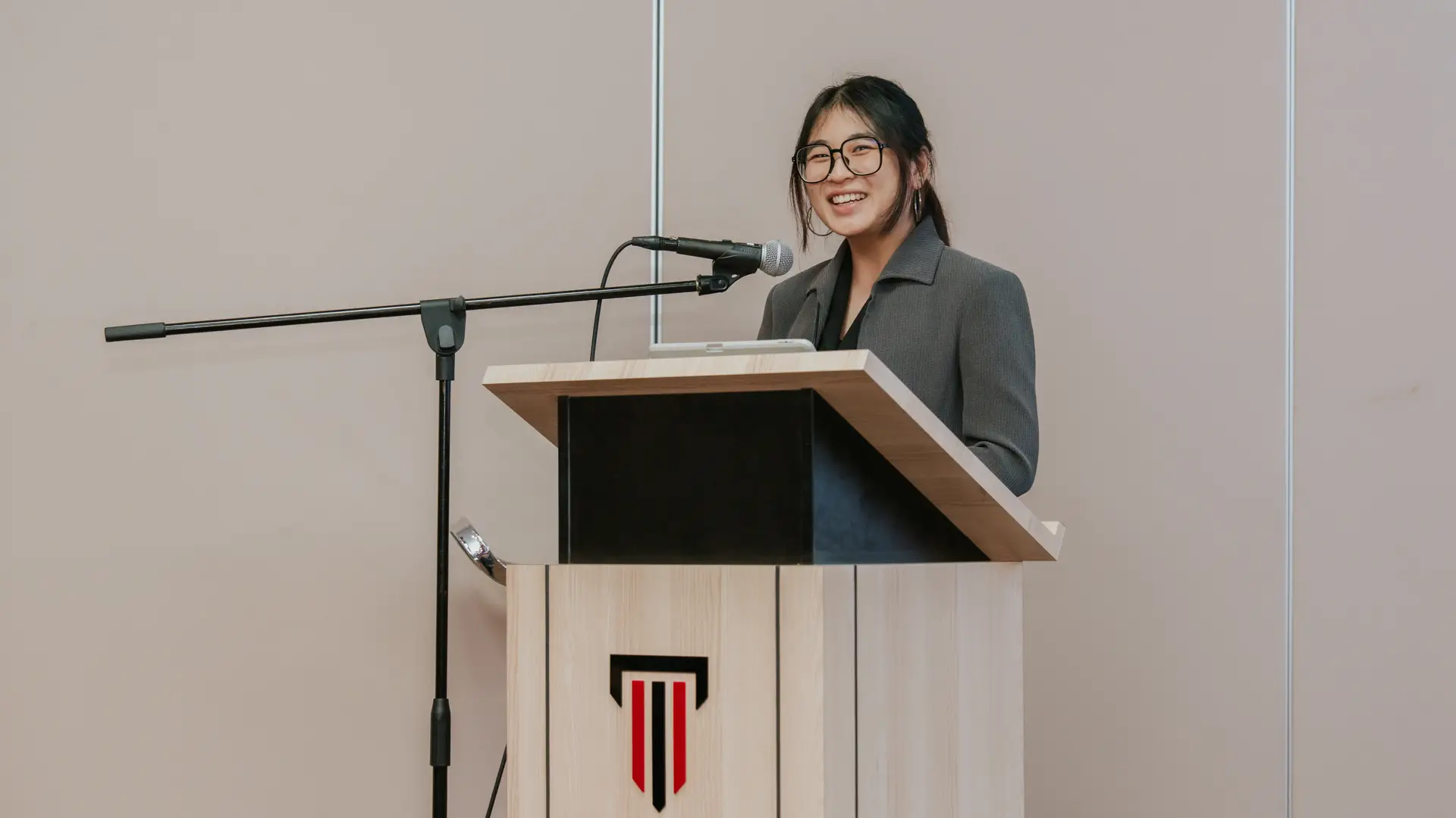 A Taylor's student in a gray blazer speaking at a podium with a microphone, smiling during a presentation.