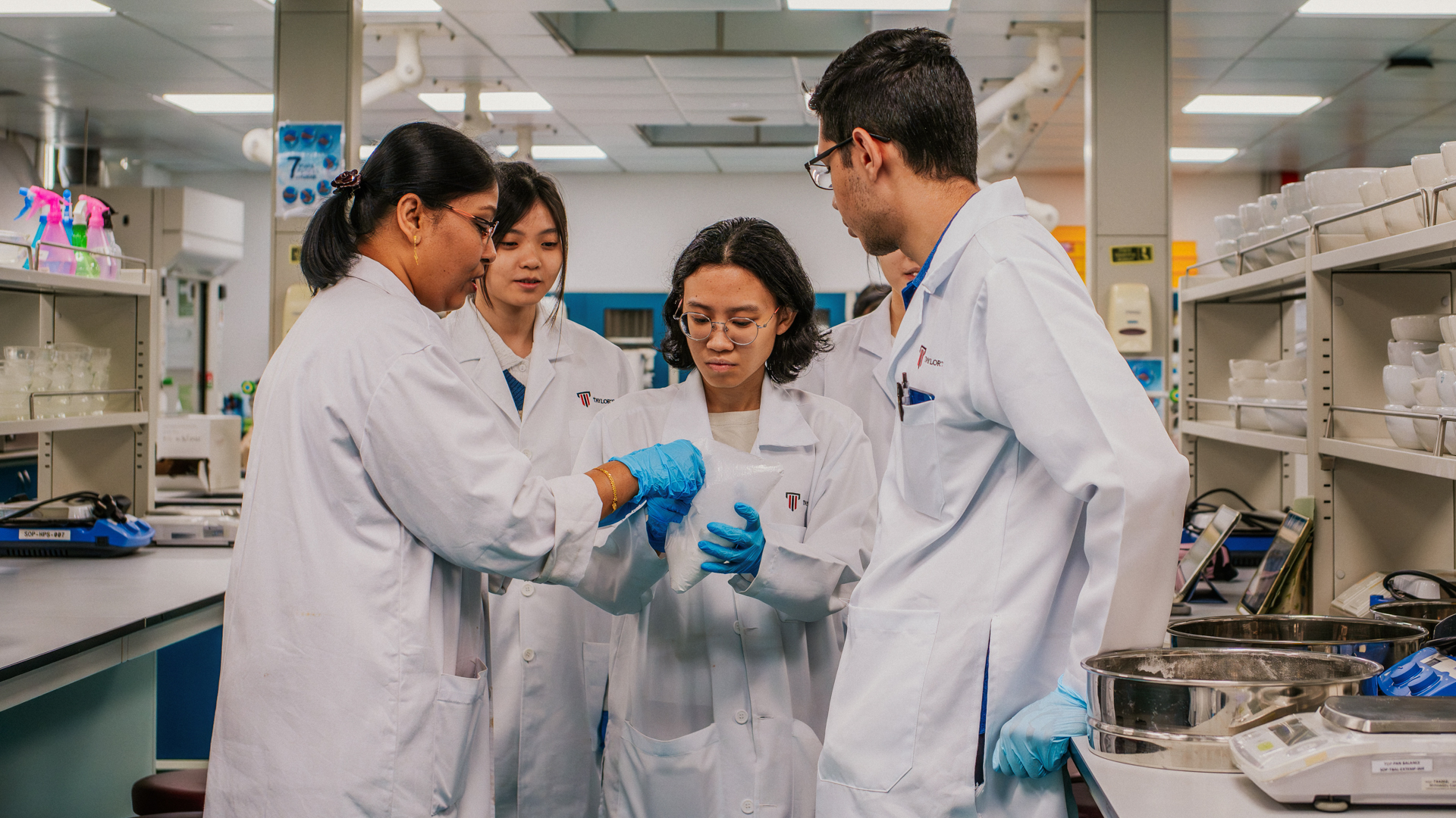 Taylor's pharmacy students in lab coats and gloves examining a bag of white powder together in a laboratory.
