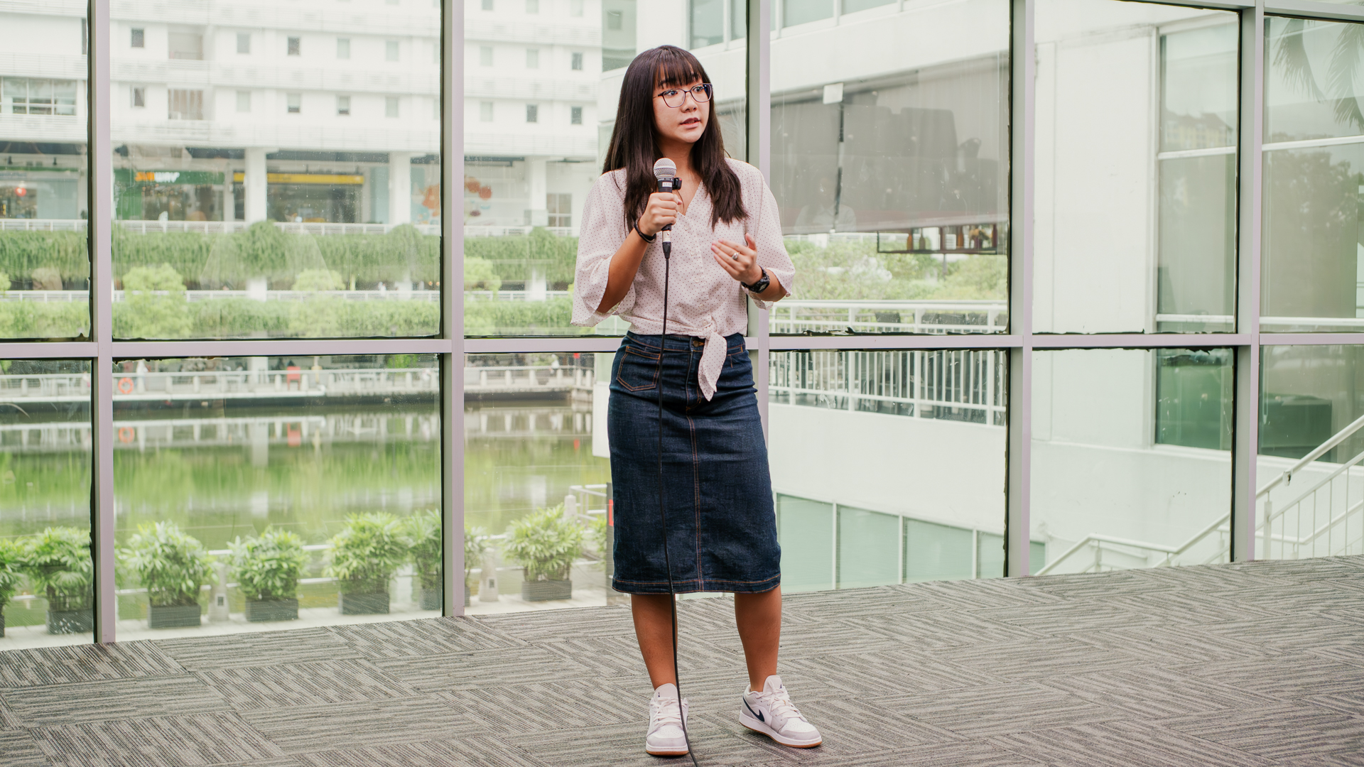 A Taylor's student stands on a stage holding a microphone and giving a presentation, with large floor-to-ceiling windows behind them overlooking a landscaped outdoor area.