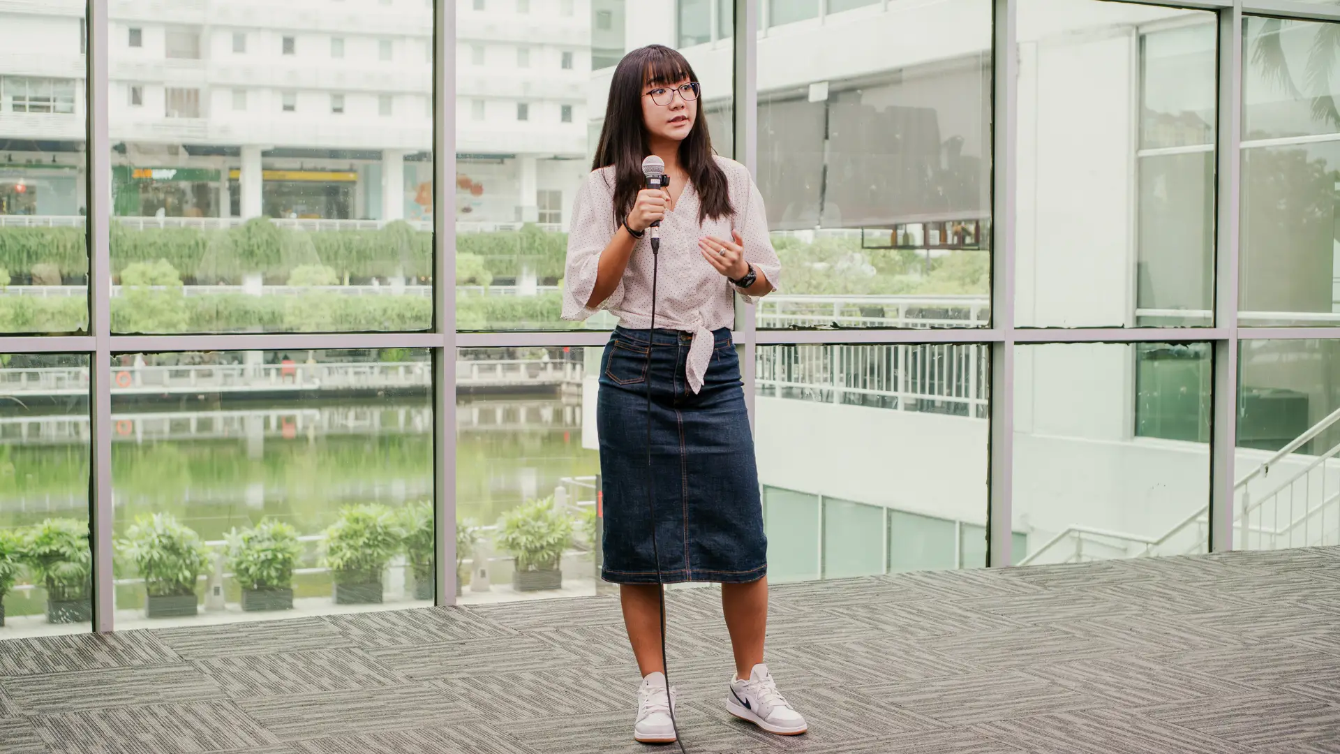 A Taylor's student stands on a stage holding a microphone and giving a presentation, with large floor-to-ceiling windows behind them overlooking a landscaped outdoor area.